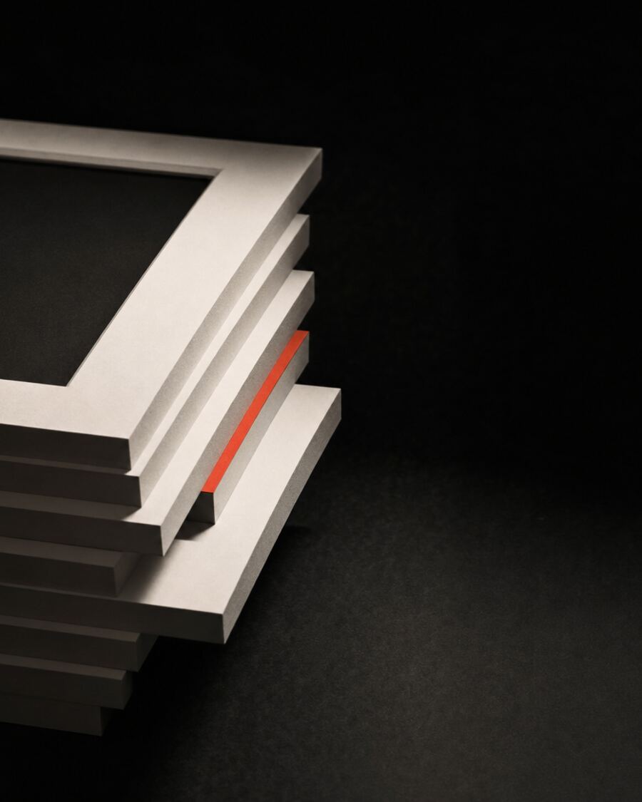 A stack of blank white paper frames on a dark studio bench, each offset from the one below, one pulled clearly out of alignment, with a thin strip of red tucked between two of them.