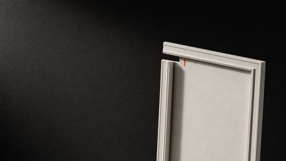 A blank white frame standing against a dark wall, a signal-orange tab marking its top edge and a small offcut resting beside it.